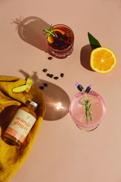 Two cocktails with herbs, orange slice, ginger, coffee beans, and orange ginger syrup bottle.