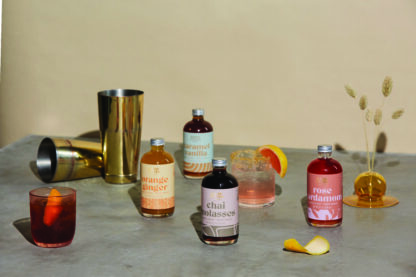 Bottles of flavored syrups and cocktails on a gray surface with gold shakers and decor.